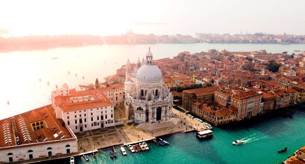 Aerial view of a historic waterfront city with domed building, red-tiled roofs, and turquoise water under soft warm light